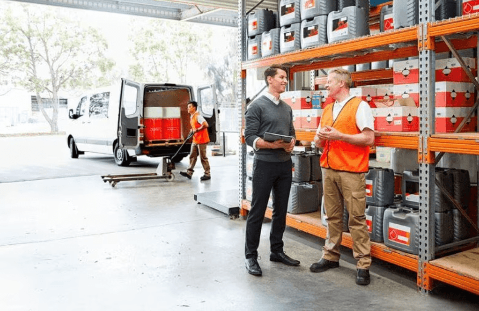 Warehouse staff loading a delivery van at a loading dock while two colleagues talk beside the racking
